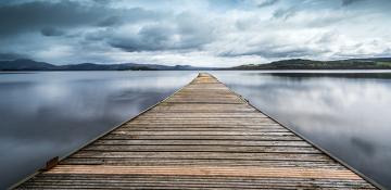 image of Scottish Loch Lomond. Features a wooden deck stretching out over the water. With a grey and moody sky over rolling hills.