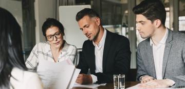 Image shows three people in business attire looking at documents and talking. Sat around the table the men are wearing suits and jackets and the women is wearing a shirt.