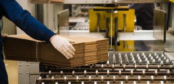 Image shows packaging line for flat pack cardboard boxes. A man wearing a jumpsuit and protective gloves is moving them from a conveyor belt that is tying stacks of boxes together