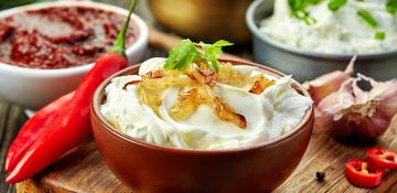 Image shows several small bowls with different dips, yogurt and chilli. It has items of food used in the preparation of the dips on chopping boards including garlic and chilli