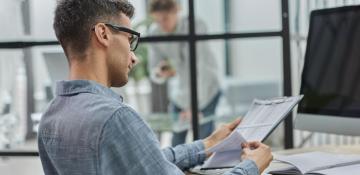 Image shows man wearing glasses, sat a a desk in front of a computer. He is wearing a shirt and looks to be in an office. With a clipboard in hands he is looking through papers.
