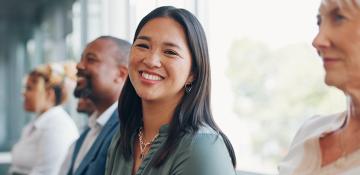 Image shows Aisan women, with long dark hair and wear a green shirt, looking at the camera and smiling. She is in a corporate office in a meeting, with co-workers either side of her. 