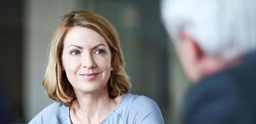 Image shows blonde white woman in focus of shot. To the right of her, out of focus is a person with short grey hair.