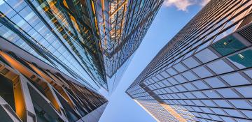 Image shows high rise glass buildings in a city centre. The perspective is from below looking up at the sky and the tops of the buildings.