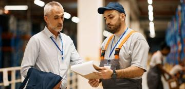 Two men are standing in a warehouse. One man is wearing a white shirt and holding a jacket, while the other man is dressed in work overalls with orange suspenders and holding a clipboard. Both men have lanyards with identification badges around their necks. The background shows shelves stocked with items and another worker handling boxes.