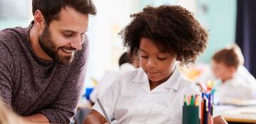 A teacher in a maroon sweater sits next to a student in a white school uniform in a classroom. Other students are working at their desks in the background, with school supplies like colored pencils and books on the desk in the foreground.