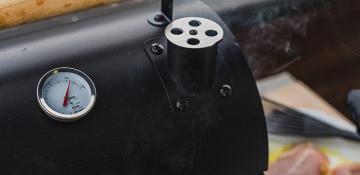Close-up of a black metal smoker with a temperature gauge reading 225°F and a cylindrical vent on top, with smoke rising and blurred grilling tools in the background.