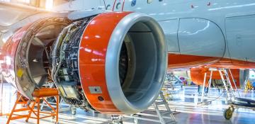 Close-up view of an aircraft engine undergoing maintenance in a hangar. The engine's red outer casing is partially removed, exposing intricate internal components and wiring. Surrounding the engine are tools, ladders, and stands, with parts of the airplane's fuselage and other maintenance equipment visible in the background.